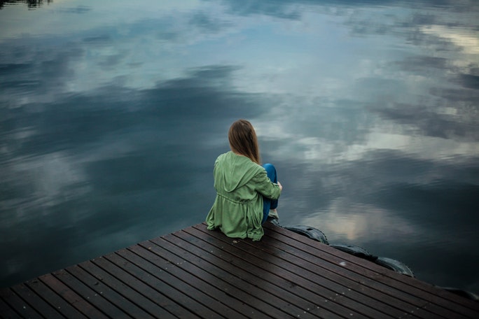 Soziale Phobie Test: Mädchen mit Angststörung sitzt auf einem Steg am Ufer eines düsteren Sees, die Knie mit den Armen umschlungen